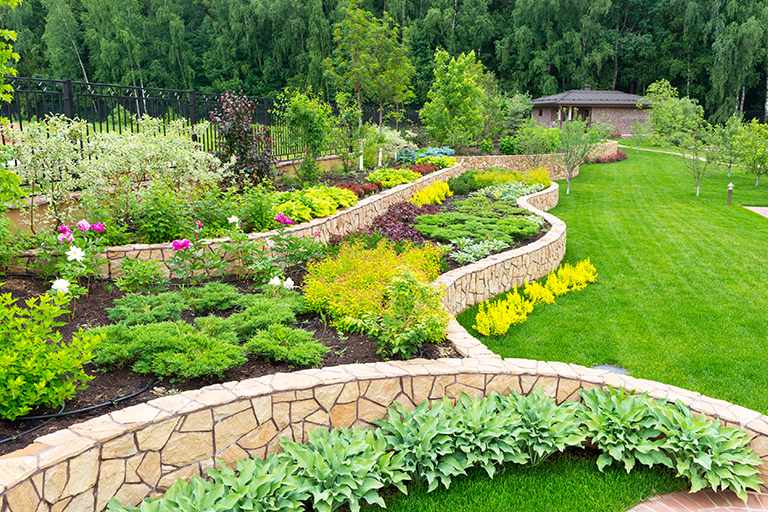 Backyard garden with flowers, trees, green grass, and rock retaining wall | Consider Starting Your New Backyard Landscape Design Ideas This Winter | Hittle Landscape